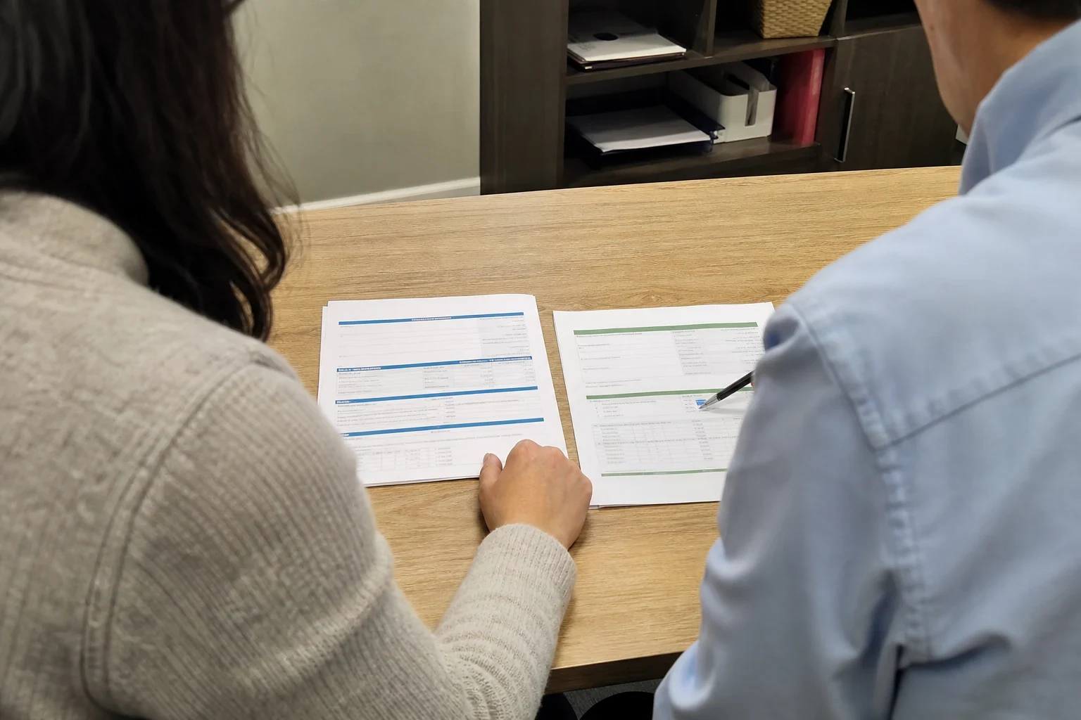 Two people seen from behind at a wooden office desk reviewing two printed documents side by side, one pointing at a table of figures with a pen.