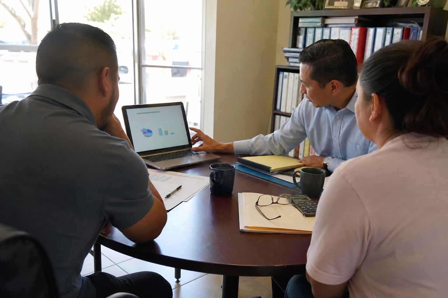 Agent in a light blue button-down points to charts on a laptop while a couple sits at a round table in a storefront office with binders on shelves