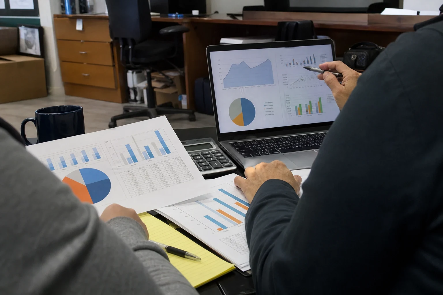 Over-the-shoulder view of two people reviewing printed charts, a laptop showing graphs, a calculator and a notepad on a desk in a small back office